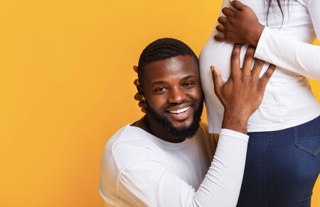 Emotional african man listening to his pregnant wife belly over yellow studio background, close up, free spaceの写真素材