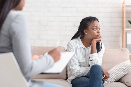 Thoughtful black woman sitting on couch at psychotherapists office, looking indifferently at window, not ready to share her problems, copy spaceの写真素材