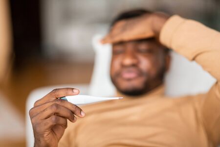 Fever. Sick Black Guy Measuring Body Temperature With Thermometer Lying On Couch At Home. Selective...