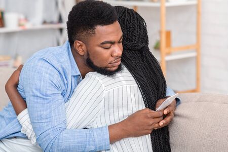 Cheating husband. Young african american man texting on cellphone with somebody while embracing his wife at home, closeupの写真素材