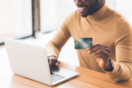Online Shopping Concept. Happy african guy holding plastic credit card and using laptop. Copy spaceの写真素材