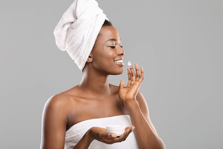 Skincare routine. Pretty african american girl with perfect skin applying moisturising cream to her cheek after bath, grey background with copy spaceの写真素材