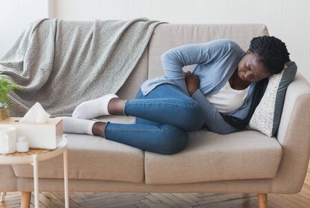 Menstrual Pain. Sick Young Black Woman Lying On Couch At Home Suffering From Abdominal Acheの写真素材
