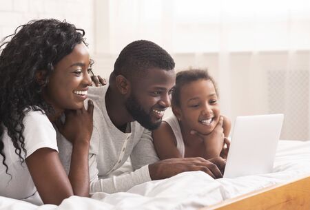 Joyful black family of three using laptop in bed together, browsing internet or watching movieの写真素材