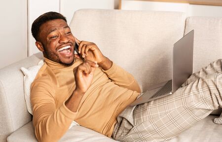 Cheerful Black Guy Talking On Phone With Friend And Laughing Lying With Laptop On Couch At Home. Modern Communicationの写真素材