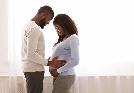 Loving afro man holding his wife belly over big window, parenthood concept, copy spaceの写真素材