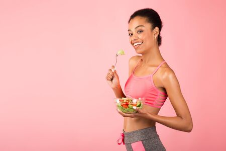 Workout And Nutrition. Black Girl Eating Vegetable Salad From Bowl Standing On Pink Studio Background. Copy Spaceの写真素材