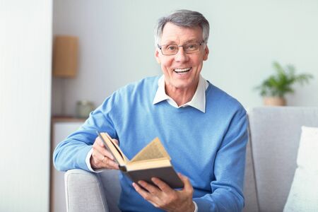 Business Literature. Elderly Man Reading Book Smiling At Camera Sitting On Couch At Home. Selective Focusの写真素材