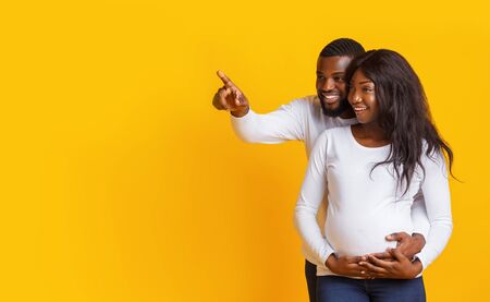 Excited african husband showing beautiful pregnant wife amazing sale, pointing at copy space over...