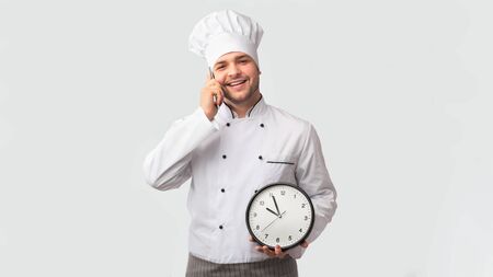 Culinary Services. Smiling Chef Cook Man In Uniform Talking On Phone Holding Clock Standing On White Studio Background. Panoramaの写真素材