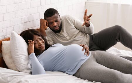 Pregnant afro couple fighting in bedroom at home home, crying pregnant woman and nervous man laying on bedの写真素材