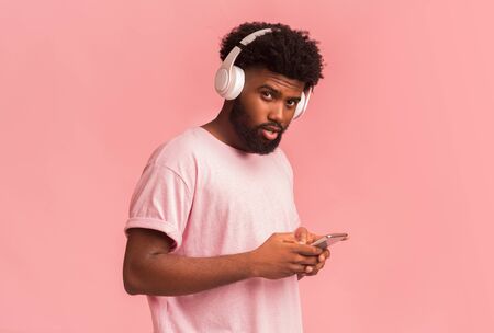 Nice music app for smartphone. Young black man with wireless headset using phone over pink backgroundの写真素材
