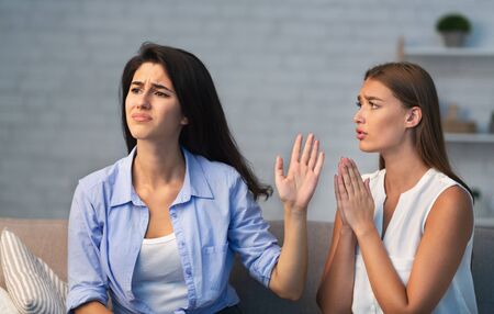 Miserable Girl Asking Indifferent Friend For A Favor Begging Holding Hands In Prayer Gesture Sitting On Sofa Indoor. Selective Focusの写真素材