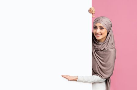 Look here. Beautiful muslim girl in hijab pointing at white blank advertisement board for your text with open palm, pink studio backgroundの写真素材