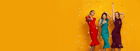 Dance Party. Three Happy Girls In Elegant Dresses Having Fun With Falling Silver Confetti On Yellow...