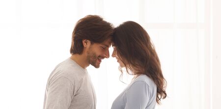 Romantic young couple in love touching foreheads, bonding to each other against window background,...