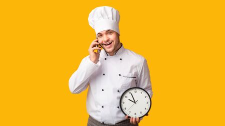 Catering Food. Chef Man Talking On Cellphone Holding Clock Standing Over Yellow Studio Background. Panoramaの写真素材