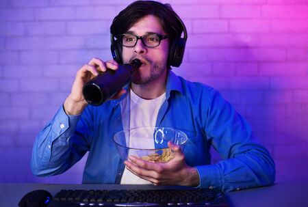 Concentrated gamer drinking beer with chips, playing games online and looking at screenの写真素材