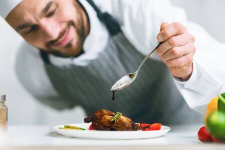 Happy Chef Man In Uniform Cooking Food Pouring Sauce On Chicken Dish Standing In Restaurant Kitchen...
