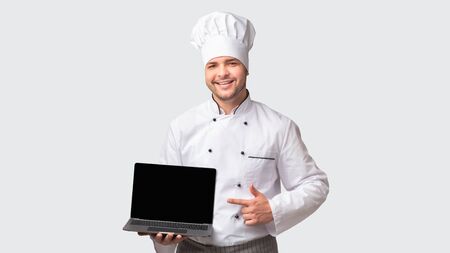 Chef Man Holding Laptop With Blank Screen Pointing Finger Standing Over White Studio Background. Cooking And Recipes Website. Panoramaの写真素材
