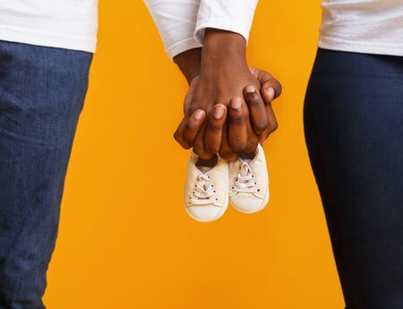 Cropped of afro couple holding baby boots together over yellow studio background, baby is coming