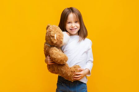 Smiling beautiful little girl embracing big teddy bear over yellow background, kids toys conceptの写真素材