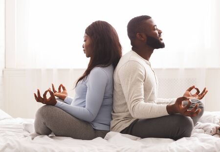 Prenatal yoga. Expecting black woman doing yoga with her husband at home, sitting on back to back with closed eyesの写真素材