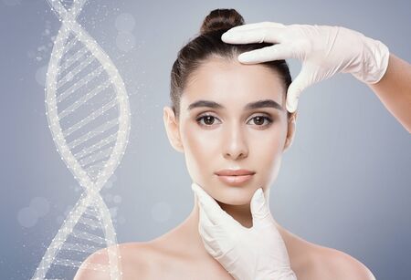 Beautiful young woman next to DNA chain over grey background, touched by cosmetologist in white...