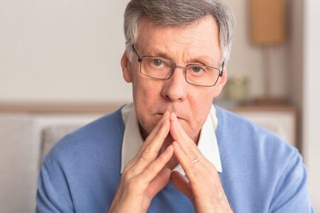 Portrait Of Serious Senior Man Thinking About Retired Life Holding Hands Near Face Sitting On Sofa Indoor. Selective Focusの写真素材