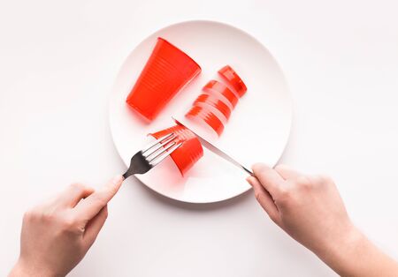 Microplastic in food. Man eating sausage of red plastic from plate, isolated on white background