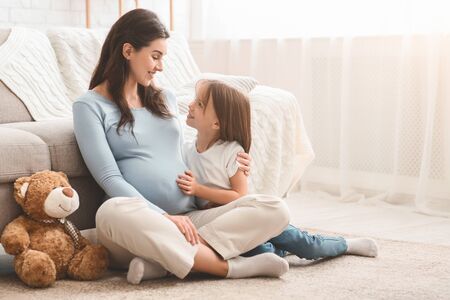 Pregnant mother and her happy daughter playing together, sitting on floor at home. Family love, motherhood and care concept, copy spaceの写真素材