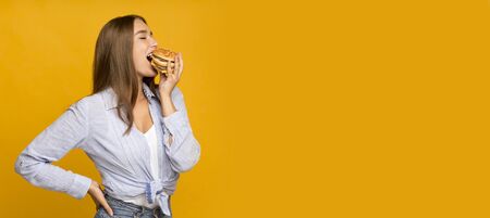Cheat meal. Hungry millennial girl eating burger enjoying unhealthy fast food standing on yellow...