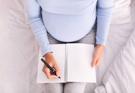 Pregnancy Diary Book. Unrecognizable Pregnant Woman Holding Notebook Taking Notes Sitting On Bed...