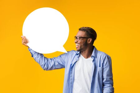 Opinion. Happy Afro Guy Holding Blank White Speech Bubble Showing What He Thinks Standing Over Yellow Studio Background. Mockupの写真素材