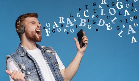 Millennial guy in wireless headphones listening music on smartphone and singing with diverse alphabet letters coming out of his mouth, blue backgroundの写真素材