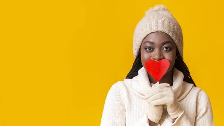 Lovely african winter woman covering her mouth with red heart shaped card over yellow background, panorama with free spaceの写真素材