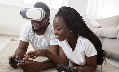 Cheerful african american couple testing VR glasses while playing video games at home together,...