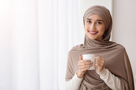 Relaxed arabic woman in headscarf standing with cup of coffee near window at home and looking at camera, enjoying weekend, empty spaceの写真素材