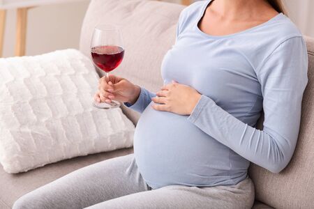 Alcohol during pregnancy. Unrecognizable mother-to-be holding glass of red wine sitting on sofa indoor. Croppedの写真素材