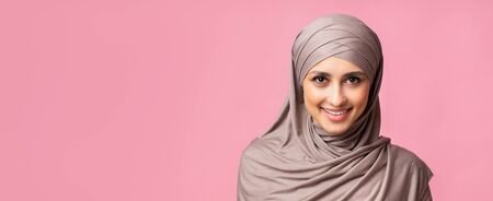 Portrait of confident muslim woman in hijab smiling and looking at camera, pink studio background,...