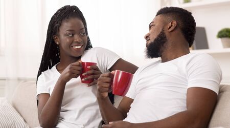 Young african american couple relaxing at home, drinking coffee and chatting, sitting on sofa in living room, panorama with free spaceの写真素材