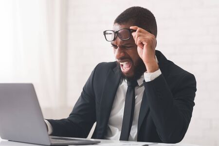 Crisis at job. Angry black entrepreneur looking at laptop screen and shouting, office interior, copy spaceの写真素材