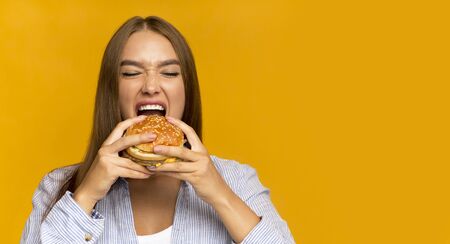 Young Lady Biting Burger Enjoying Junk Food On Cheat Meal Day Standing Over Yellow Background. Panorama, Studio Shot, Copy Spaceの写真素材