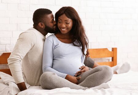 Cheerful young pregnant african couple cuddling in bed at home, waiting for baby, copy spaceの写真素材