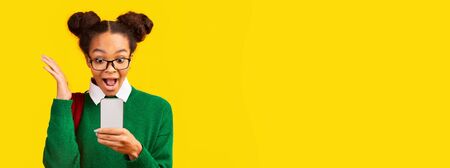 Pleasant Surprise. Shocked black teen girl looking at smartphone over yellow background, panorama, copy spaceの写真素材