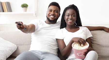 Happy afro couple watching tv and eating popcorn, relaxing on sofa at home, enjoying weekend together, panoramaの写真素材