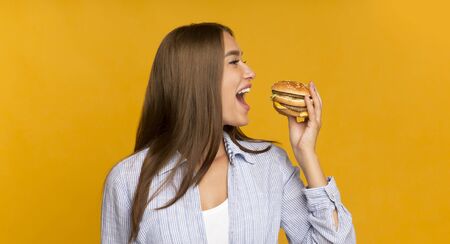 Junk Food. Happy Girl Eating Tasty Unhealthy Burger Standing On Yellow Background. Studio Shot,...