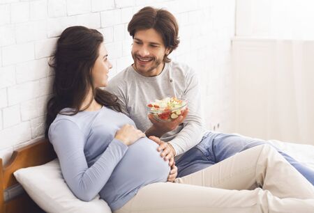 Healthy Nutrition During Pregnancy Concept. Loving husband giving fresh vegetable salad to his...