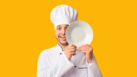 Funny Cook Man Covering Eye With Plate Looking At Camera Standing Over Yellow Studio Background. Panoramaの写真素材