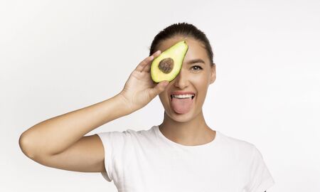 Playful Woman Holding Avocado Half Covering Eye And Showing Tongue Having Fun Over White Studio Background.の写真素材